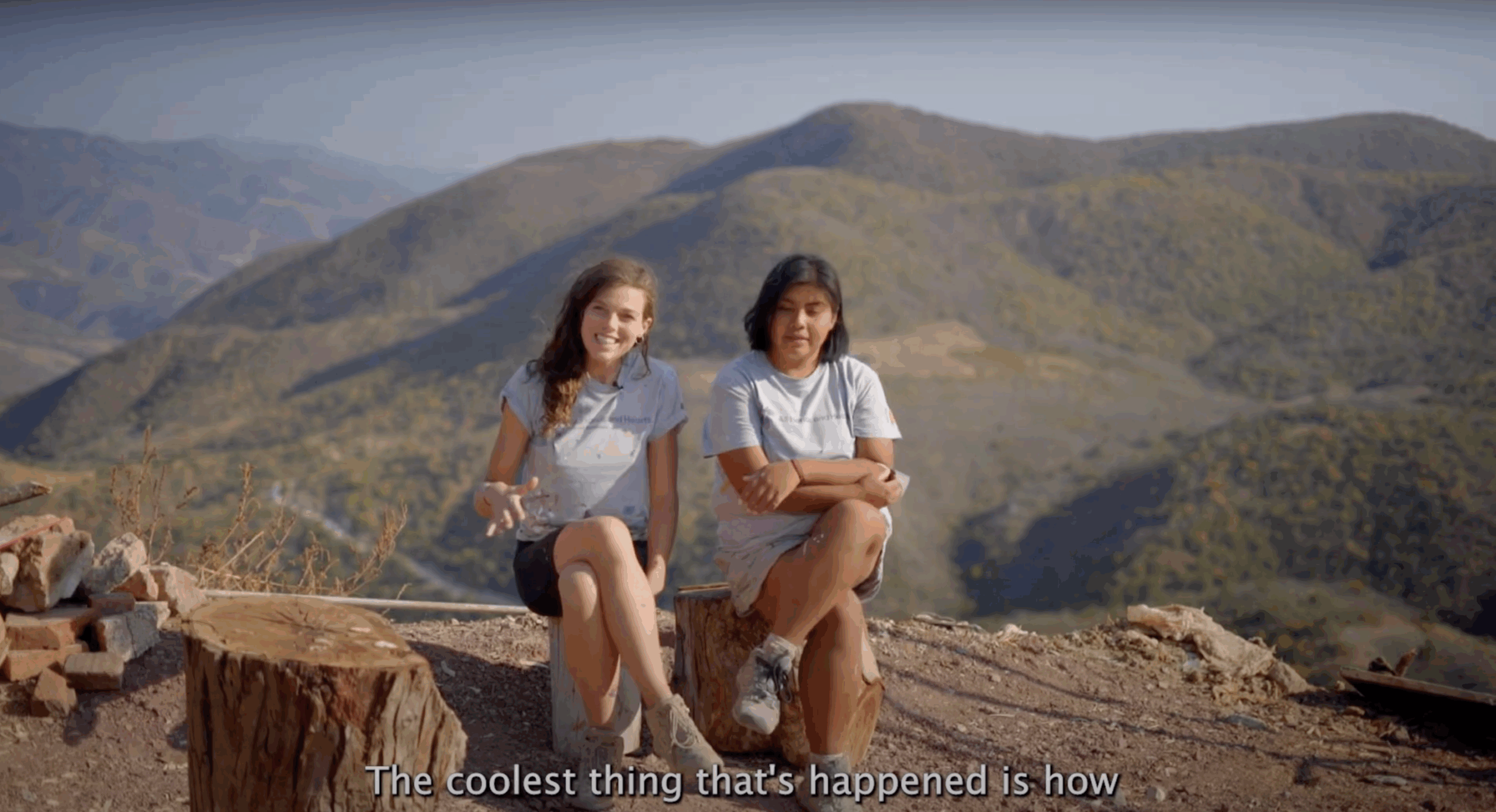 Two young women sit on tree stumps overlooking a vast mountain landscape. They are wearing light gray "All Hands and Hearts" t-shirts and work boots, smiling and relaxed. One gestures while speaking, as the other sits with arms crossed. Subtitles at the bottom of the image read, “The coolest thing that’s happened is how.”